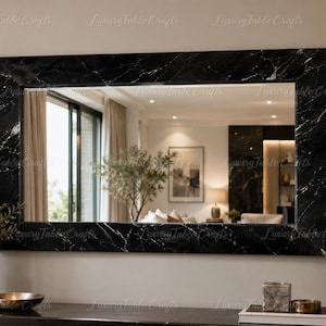 May include: A rectangular wall mirror featuring a wide, polished black marble frame with prominent white veining. The mirror surface reflects a room interior, including a potted plant, a framed picture, and a lamp, with a watermark text overlay.