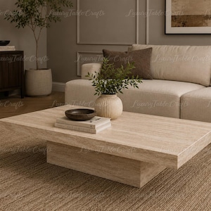 Pode incluir: Uma mesa de centro retangular bege claro com uma base sólida, uma pequena tigela de madeira e uma pilha de livros. A mesa está numa sala de estar com um sofá bege e um tapete castanho.