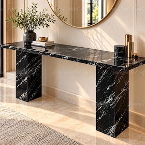 Può includere: Una lunga console rettangolare con superficie in marmo nero venato di bianco, sostenuta da due spesse gambe in marmo abbinate. Il tavolo ospita un vaso scuro con vegetazione, una piccola ciotola su libri e due portacandele cilindrici dorati.
