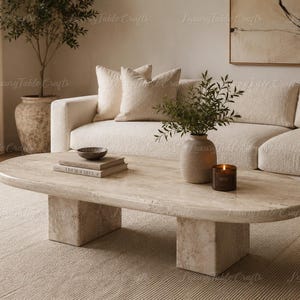 May include: An oval coffee table made of beige travertine stone with a textured surface and two thick, rectangular block legs. The table is positioned in a living space, supporting a small dark bowl, two stacked books, a vase, and a candle.