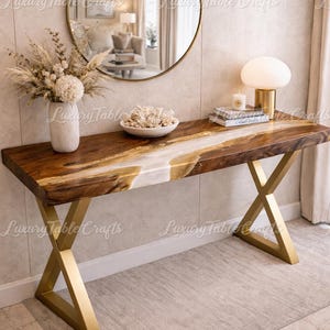 Op de afbeelding: Een moderne console met een houten blad met een natuurlijke rand en goudkleurige metalen poten. De tafel heeft een wit en goud hars rivierontwerp. Een ronde spiegel en decoratieve items staan op de tafel.