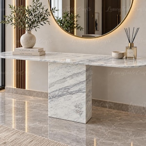 May include: A white marble console table with a long, oval-shaped top and a single rectangular pedestal base. The surface features grey veining throughout. The table is positioned in a room with a large round mirror and decorative items on top.