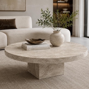 May include: A round, beige travertine coffee table with a thick, cylindrical top and a square pedestal base. On the table sits a beige stone vase with green foliage, a dark brown wavy bowl, and two stacked books with the word Kinfolk visible.