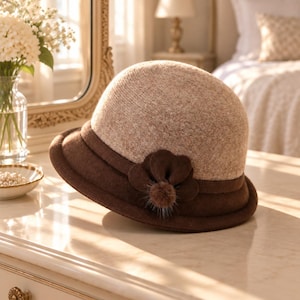 May include: A two-tone cloche hat featuring a textured beige crown and a solid dark brown brim. The hat is accented with a dark brown fabric flower detail on the side, which includes a small, fuzzy pom-pom center, creating a classic rounded silhouette.