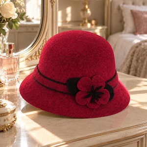Puede incluir: Un sombrero tipo cloche de fieltro rojo intenso con una copa redondeada y un ala ligeramente inclinada hacia abajo. El sombrero está adornado con una fina banda negra y un detalle de flor de tela a juego con pétalos centrales negros y hojas verde oscuro.