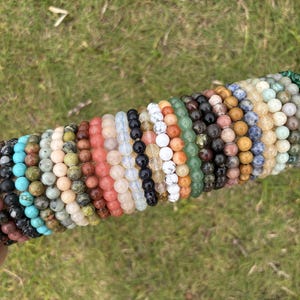 May include: A selection of beaded bracelets in diverse colours, including brown, turquoise, black, white, green, and pink. The bracelets are made of natural stones and are displayed on a tan cylindrical surface against a backdrop of green grass.