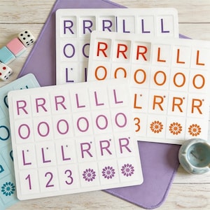 Könnte beinhalten: Weiße Spielsteine mit lila, orangefarbenen und roten Buchstaben und Zahlen. Die Steine sind auf einer lila Matte angeordnet. Würfel und andere Spielsteine sind im Hintergrund zu sehen. Die Spielsteine haben die Buchstaben R, L und O.
