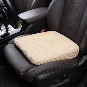 Puede incluir: Un cojín de asiento de coche cuadrado beige con una superficie texturizada. El cojín está colocado sobre un asiento de coche de cuero marrón oscuro. El interior del coche incluye un volante y una consola central.
