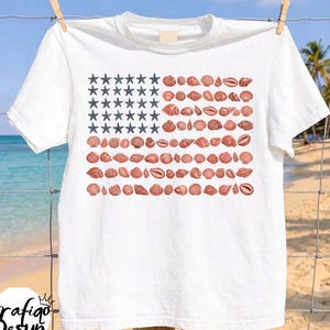 Puede incluir: Una camiseta blanca con un diseño gráfico de la bandera estadounidense compuesto por estrellas de mar azul oscuro en el cantón y filas de varias conchas marinas marrones que forman las franjas. La camiseta cuelga de una percha de madera.
