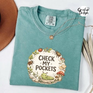 Peut inclure: T-shirt de couleur sarcelle avec l'inscription "CHECK MY POCKETS" entourée d'une couronne de feuilles, d'insectes et de champignons. Un collier argenté avec un pendentif est posé sur le t-shirt. Le t-shirt est en matière Comfort Colors.