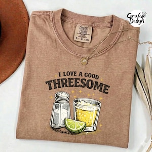 May include: A brown Comfort Colors t-shirt with the phrase "I LOVE A GOOD THREESOME" printed on the front. The graphic includes a salt shaker, a glass of beer, and a lime wedge. A gold necklace is draped across the shirt.
