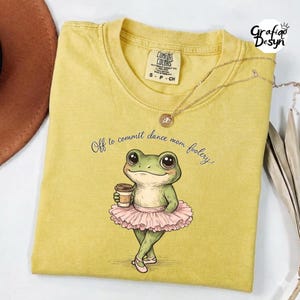Puede incluir: Camiseta de color amarillo mostaza con un gráfico de una rana bailarina sosteniendo una taza de café. La rana lleva un tutú rosa y zapatillas de ballet. El texto en la camiseta dice "Off to commit dance mom foolery."