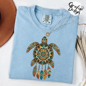 May include: A light blue crew neck t-shirt featuring a graphic of a sea turtle with a colorful mandala shell pattern and dangling feathers. The shirt has a Comfort Colors brand label at the inner neckline and a thin silver chain necklace.