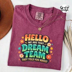 May include: A maroon Comfort Colors t-shirt with the text "HELLO WE ARE THAT DREAM TEAM THEY TOLD YOU ABOUT" in a colorful design. A gold necklace with a pendant is draped on the shirt.