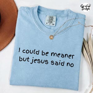 Puede incluir: Camiseta Comfort Colors azul claro con el texto negro "I could be meaner but Jesus said no". Un collar dorado con un colgante está colocado sobre la camiseta.