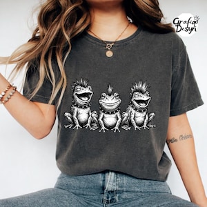 May include: Dark gray t-shirt featuring a black and white graphic of three punk rock frogs. Each frog has a mohawk, studded collar, and a unique facial expression. The shirt has a crew neck and short sleeves.