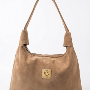 Pode incluir: Uma bolsa hobo de camurça bege com uma alça de ombro única com fixações com nós na base. A frente apresenta uma placa de metal quadrada em tom dourado com o texto OroCarry e um logótipo C estilizado. Fecho de correr na borda superior.