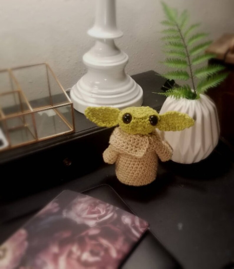 Op de afbeelding: Een gehaakt groen en beige pluche speelgoed met grote ogen, zittend op een zwart oppervlak. Het speelgoed draagt ​​een beige jas en heeft een groen hoofd en oren.