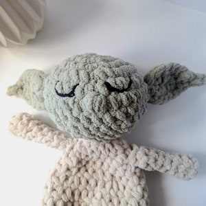 Pu&ograve; includere: Un giocattolo di peluche lavorato a uncinetto a forma di personaggio con grandi orecchie, occhi chiusi e una testa verde chiaro. Il corpo &egrave; di colore beige chiaro.