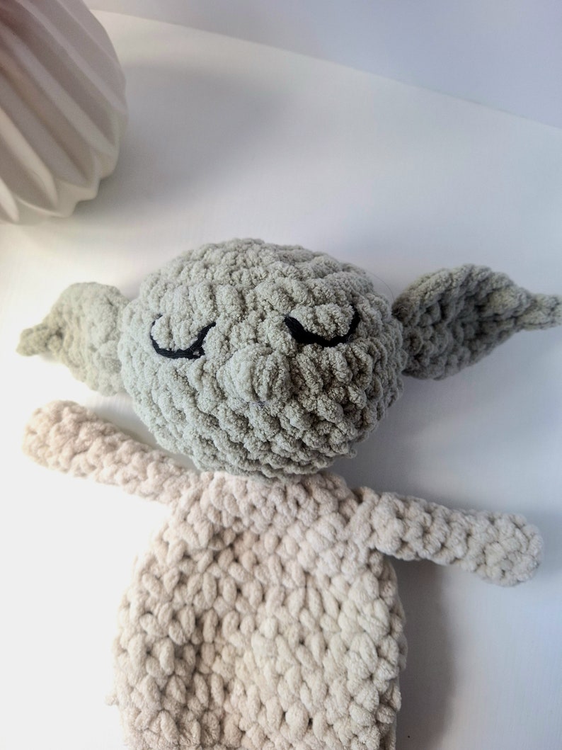 Pu&ograve; includere: Un giocattolo di peluche lavorato a uncinetto a forma di personaggio con grandi orecchie, occhi chiusi e una testa verde chiaro. Il corpo &egrave; di colore beige chiaro.