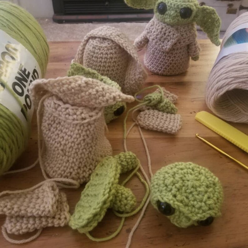 Op de afbeelding: Een verzameling gehaakte amigurumi-speelgoed in verschillende stadia van voltooiing. De speelgoed zijn groen en beige en hebben grote ogen. De speelgoed liggen op een houten oppervlak met garen en een haaknaald.