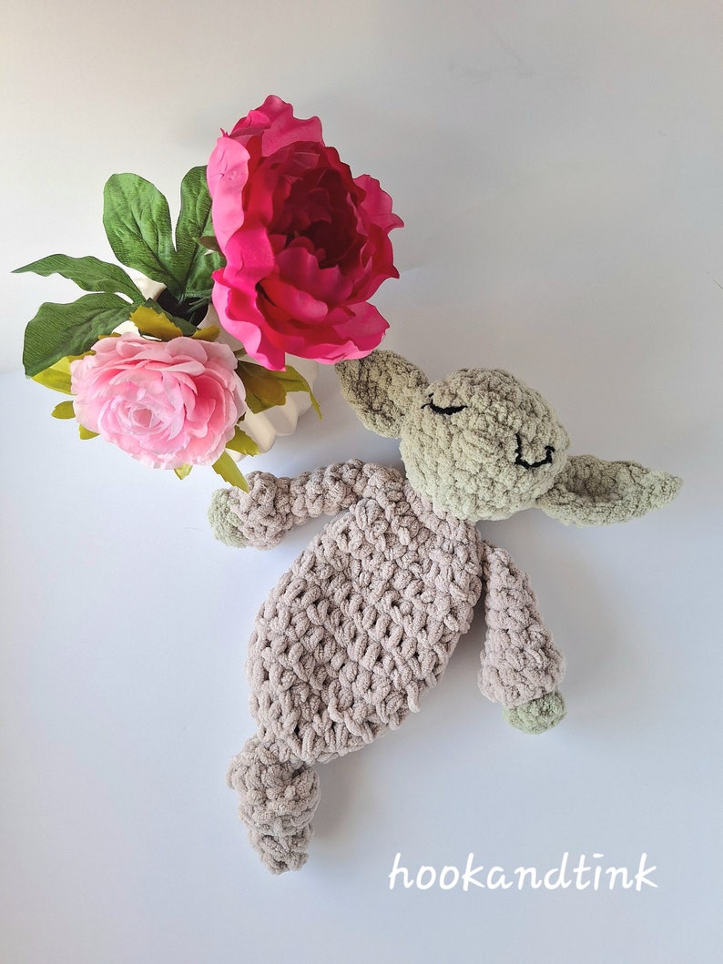 Pu&ograve; includere: Un giocattolo di peluche all'uncinetto a forma di alieno verde con un corpo marrone. Il giocattolo &egrave; sdraiato sulla schiena con le braccia e le gambe distese. Il giocattolo &egrave; circondato da fiori rosa.