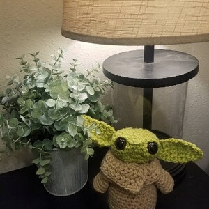 Op de afbeelding: Een gehaakt Baby Yoda speelgoed in groen en bruin met grote ogen. Het speelgoed staat op een zwart oppervlak naast een plant in een pot.