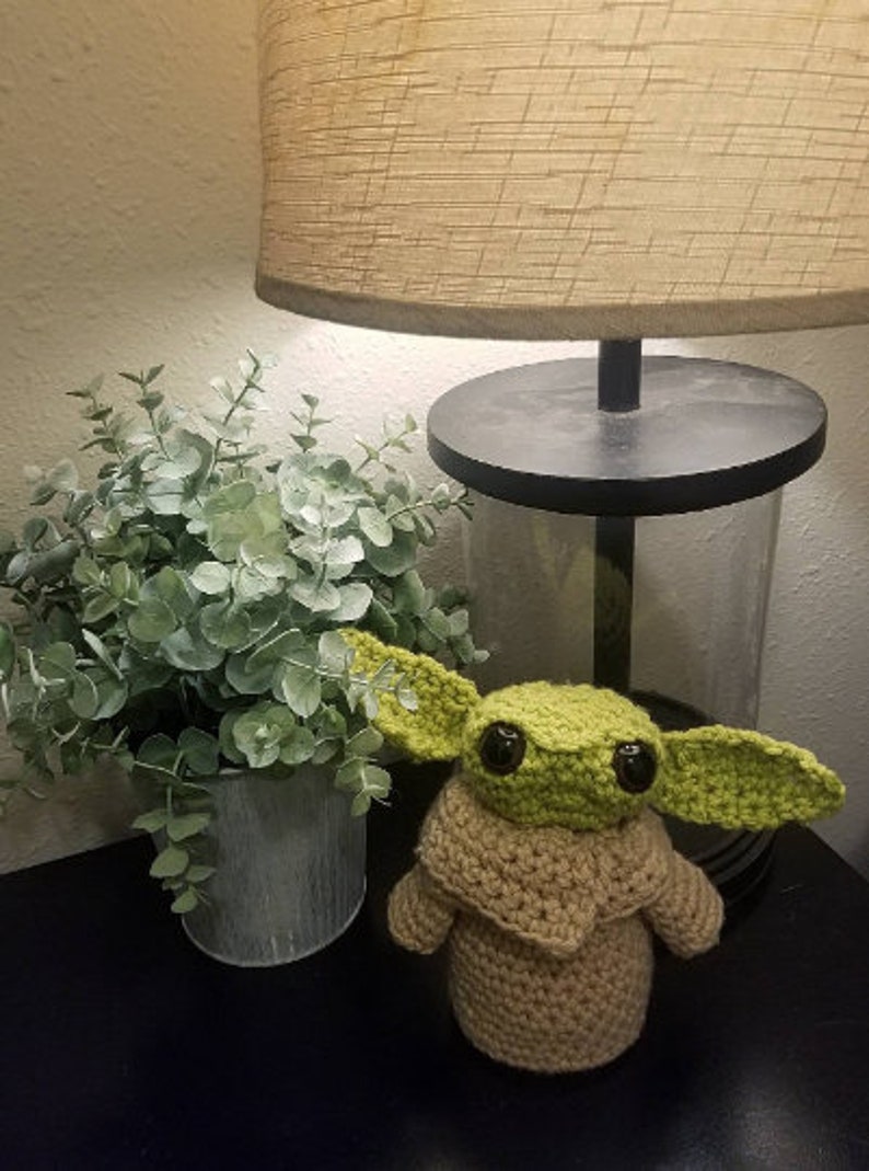 Op de afbeelding: Een gehaakt Baby Yoda speelgoed in groen en bruin met grote ogen. Het speelgoed staat op een zwart oppervlak naast een plant in een pot.