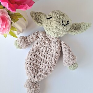 Pu&ograve; includere: Un giocattolo di peluche lavorato a uncinetto a forma di alieno verde con un corpo beige. Il giocattolo ha un viso sorridente con gli occhi chiusi ed &egrave; sdraiato su una superficie bianca.
