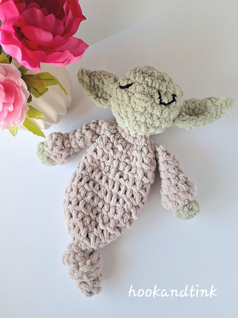 Pu&ograve; includere: Un giocattolo di peluche lavorato a uncinetto a forma di alieno verde con un corpo beige. Il giocattolo ha un viso sorridente con gli occhi chiusi ed &egrave; sdraiato su una superficie bianca.