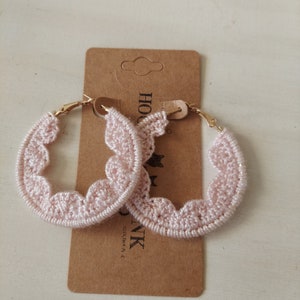 May include: A pair of pink crocheted hoop earrings with gold-toned metal hoops. The earrings are displayed on a brown card with the text "HOOK &" and "INK" printed on it.