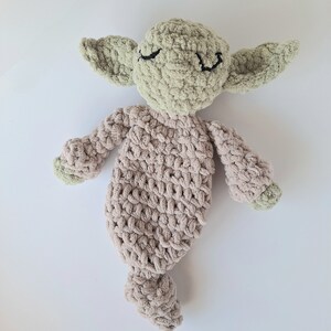 Pu&ograve; includere: Un giocattolo di peluche lavorato a uncinetto a forma di alieno verde con corpo marrone. Il giocattolo ha un'espressione con gli occhi chiusi ed &egrave; sdraiato su una superficie bianca.