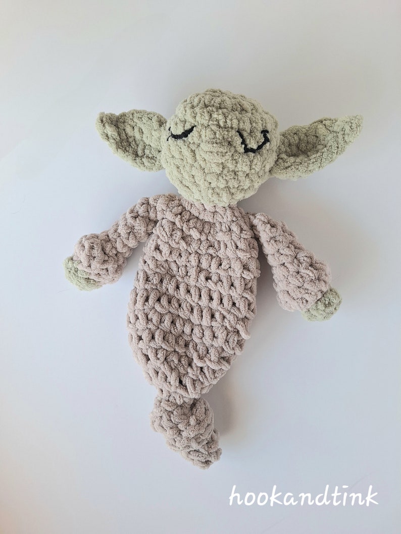 Pu&ograve; includere: Un giocattolo di peluche lavorato a uncinetto a forma di alieno verde con corpo marrone. Il giocattolo ha un'espressione con gli occhi chiusi ed &egrave; sdraiato su una superficie bianca.