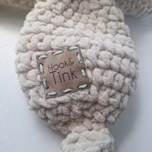 Pu&ograve; includere: Un giocattolo di peluche beige lavorato a uncinetto con un'etichetta in pelle marrone che dice "Hook & Tink".