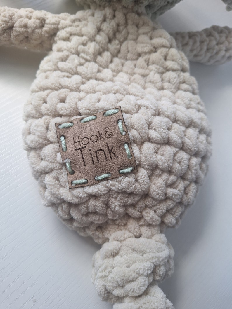 Pu&ograve; includere: Un giocattolo di peluche beige lavorato a uncinetto con un'etichetta in pelle marrone che dice "Hook & Tink".