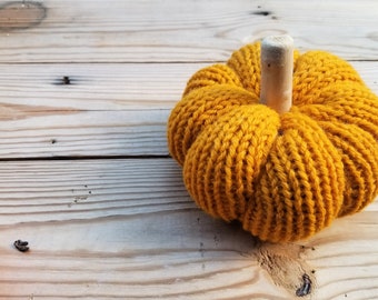 May include: A crocheted pumpkin in a mustard yellow color with a wooden stem. The pumpkin is sitting on a light brown wooden surface.