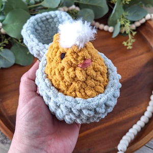 May include: A handmade crochet chick in a light blue eggshell. The chick is yellow with a white tuft of hair and a pink beak. The eggshell is open, revealing the chick inside. The item is likely a decorative Easter or spring-themed item.