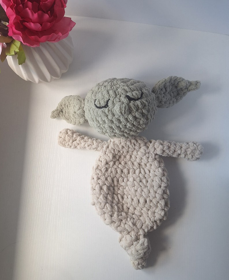 Pu&ograve; includere: Un giocattolo di peluche lavorato a uncinetto a forma di personaggio con grandi orecchie, occhi chiusi e corpo beige. Il giocattolo &egrave; realizzato con un filato morbido e peloso e ha una testa verde.