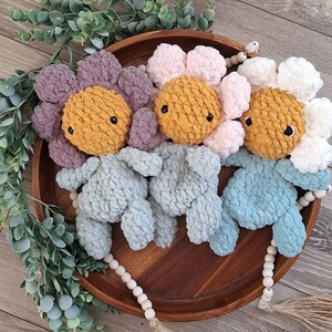 May include: Handmade crochet flower plush toys in shades of purple, pink, and white, each with a yellow center and black eyes. Three blue plush figures are below the flowers, all arranged on a wooden tray with decorative beads and greenery.