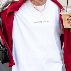 Pode incluir: Uma camiseta branca com a frase "nothing to say." é usada com uma jaqueta vermelha, boné branco e colares dourados. Uma bolsa preta está pendurada no ombro. A pessoa está segurando uma bebida de café com canudo.
