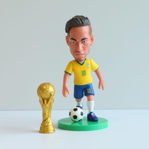 Könnte beinhalten: Figur einer Fußballspielers in gelbem Trikot mit blauen Shorts, Nummer 10, und einem Fußball auf einer grünen Basis. Eine goldene Trophäe befindet sich links von der Figur. Die Figur hat einen ernsten Gesichtsausdruck.
