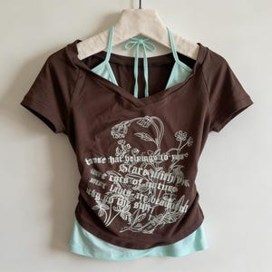 Op de afbeelding: Een bruin V-hals T-shirt met een lichtblauwe haltertop eronder. Het shirt heeft witte bloemenafbeeldingen en de tekst "those that bring us to your stars..." op de voorkant. Het shirt hangt aan een witte hanger.