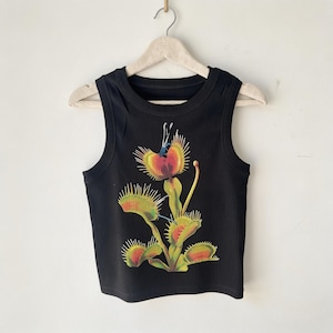 May include: A black sleeveless tank top featuring a graphic print of a Venus flytrap plant. The plant illustration displays green and reddish-orange leaves with spiky edges and a small blue insect resting on the top trap, centered on the chest.