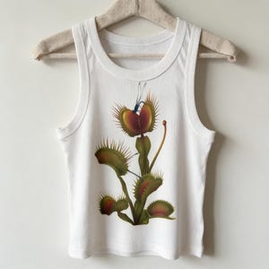 Op de afbeelding: Een wit geribbelde tanktop met een centrale grafische illustratie van een groene en rode Venusvliegenvanger met stekelige bladeren en een kleine blauwe libel die op de bovenste val zit. Het kledingstuk heeft een ronde hals en brede schouderbanden.