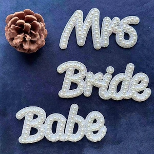 May include: White pearl-covered script words "Mrs", "Bride", and "Babe" on a dark blue background. The letters are outlined with a silver border. A brown pine cone is in the upper left corner.