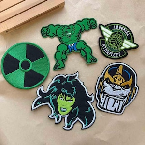 May include: A collection of embroidered patches featuring comic book and sci-fi characters. Includes a green Hulk, a She-Hulk, a Thanos, a Starfleet patch, and a green and black hazard symbol.