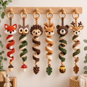May include: Six crochet animal ornaments hanging from a wooden rack. Each features a unique creature head—fox, owl, hedgehog, squirrel, bear, and deer—attached to a spiral tail with a small wooden bead and a nature-themed charm at the bottom.
