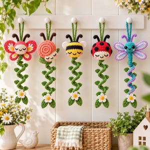 May include: Five crocheted hanging ornaments featuring a butterfly, snail, bee, ladybug, and dragonfly. Each creature has a smiling face and is attached to a green crocheted vine with a small white flower at the bottom, suspended from white hooks.