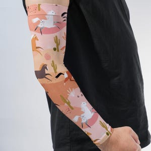 May include: An arm sleeve featuring a desert-themed pattern with various horses in motion, including white, brown, and chestnut colors. The background consists of soft pink and tan hues with green cactus illustrations and scattered brown dots.