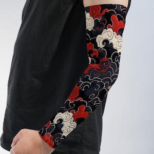 May include: A fitted arm sleeve featuring a repeating graphic pattern of stylized clouds in shades of red, cream, and black. The sleeve covers the forearm and upper arm, extending from the wrist to the bicep, worn with a plain black t-shirt.